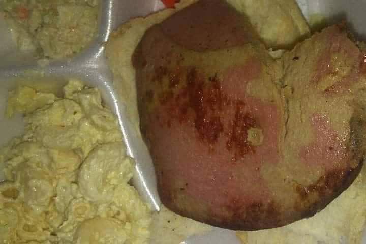 A styrofoam food tray contains what looks like scrambled eggs and an unidentified piece of food that looks like a discolored ham on it. 