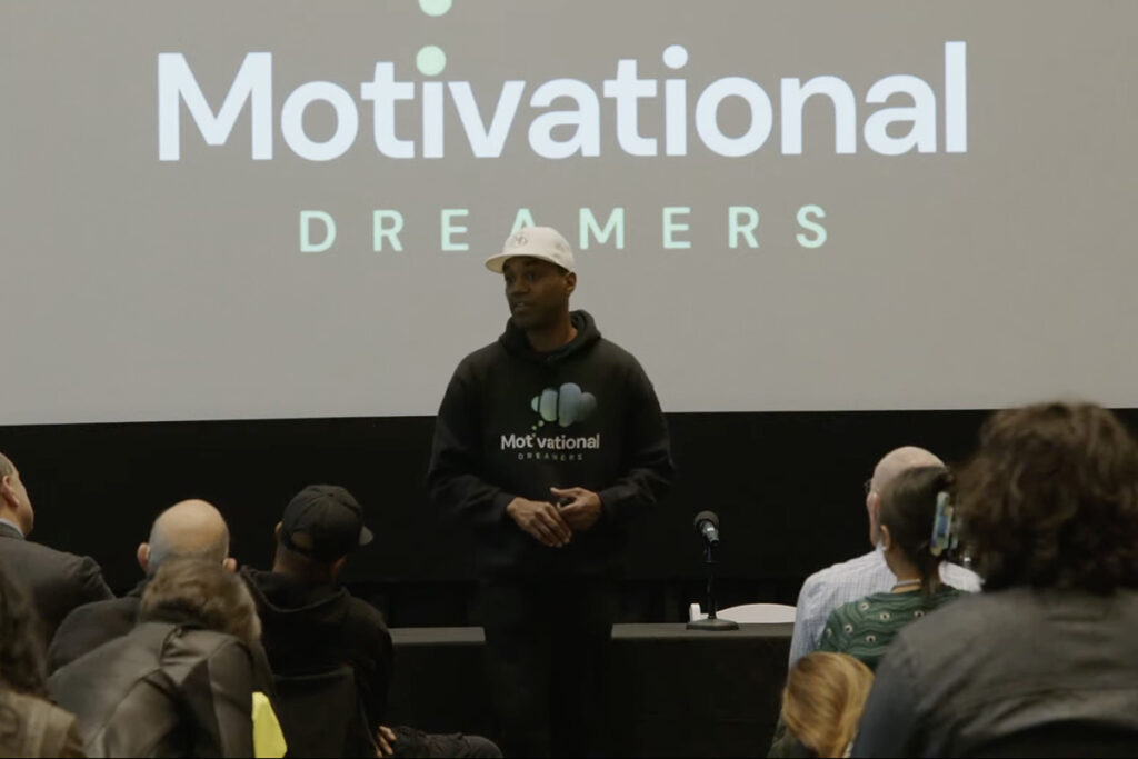 Man in black hoodie with company logo on front, white ball cap, speaks to audience in front of screen reading 