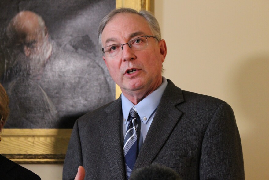 A photo of a man in a grey suit with a blue shirt and glasses, who is speaking in front of a painting of another man.
