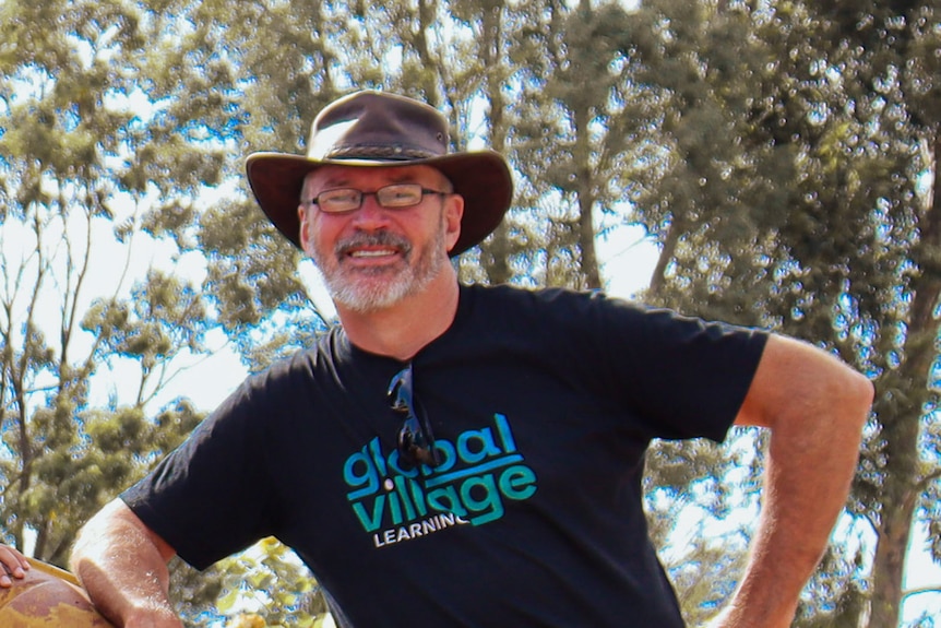 A smiling, bearded man in a hat stands in the bush on a sunny day.