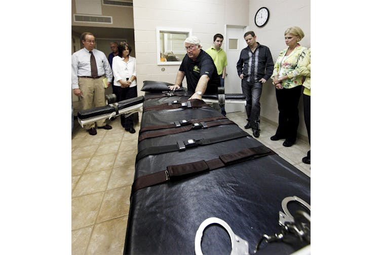 A man places his hands on a gurney in an execution chamber as five people look on.